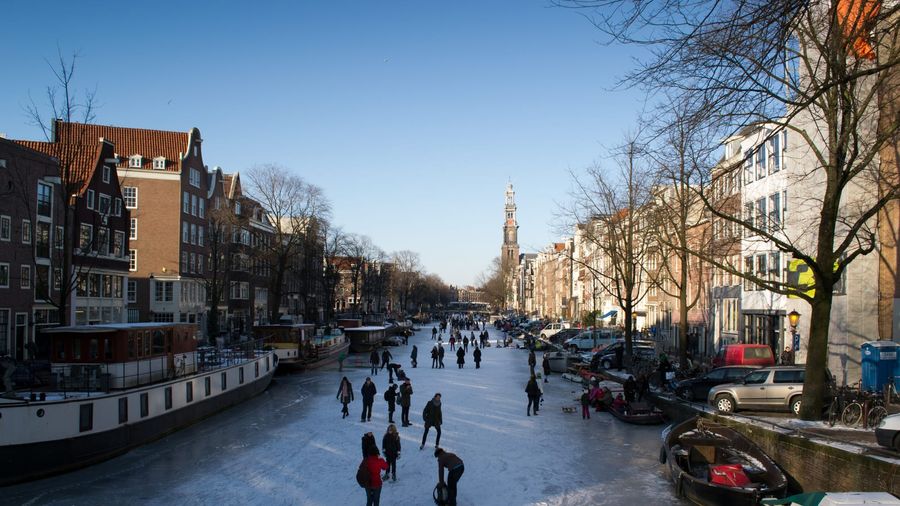 Skaters and pedestrians on a frozen Amsterdam canal lined with historic gabled canal houses and moored boats, with the Westerkerk tower rising in the distance