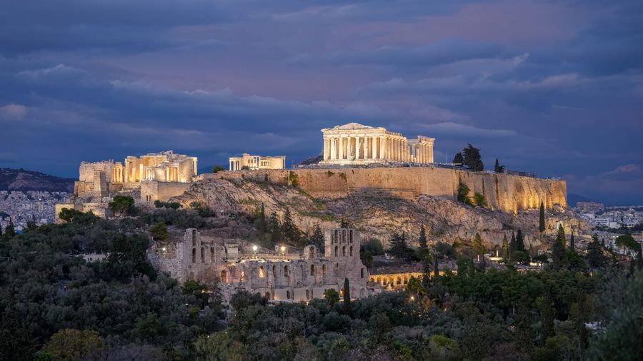 The floodlit Parthenon and ancient ruins of the Acropolis of Athens crowning a rocky, tree-covered hill above the city at twilight
