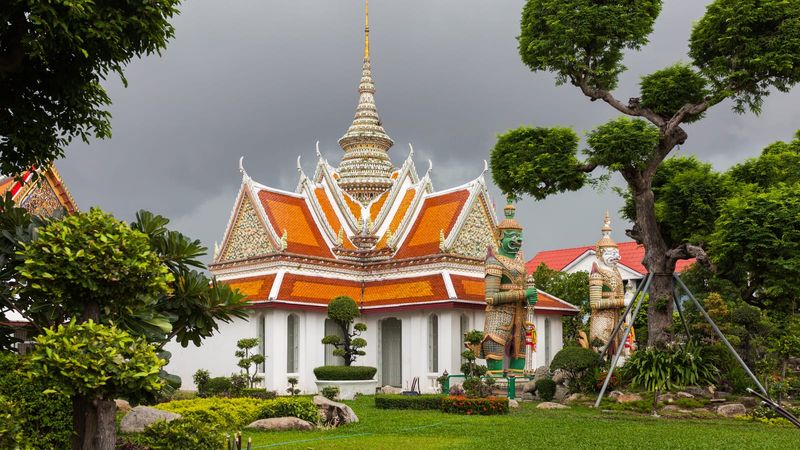 The white-and-orange tiled pavilion of Bangkok's Wat Arun temple crowned by an ornate spire, flanked by mythical guardian giant statues and manicured tropical gardens under a stormy grey sky