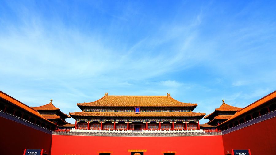 The triple-eaved golden roofs and vermilion walls of the Meridian Gate, main entrance to Beijing's Forbidden City, beneath a blue sky, with Palace Museum ticket-check signs at its base