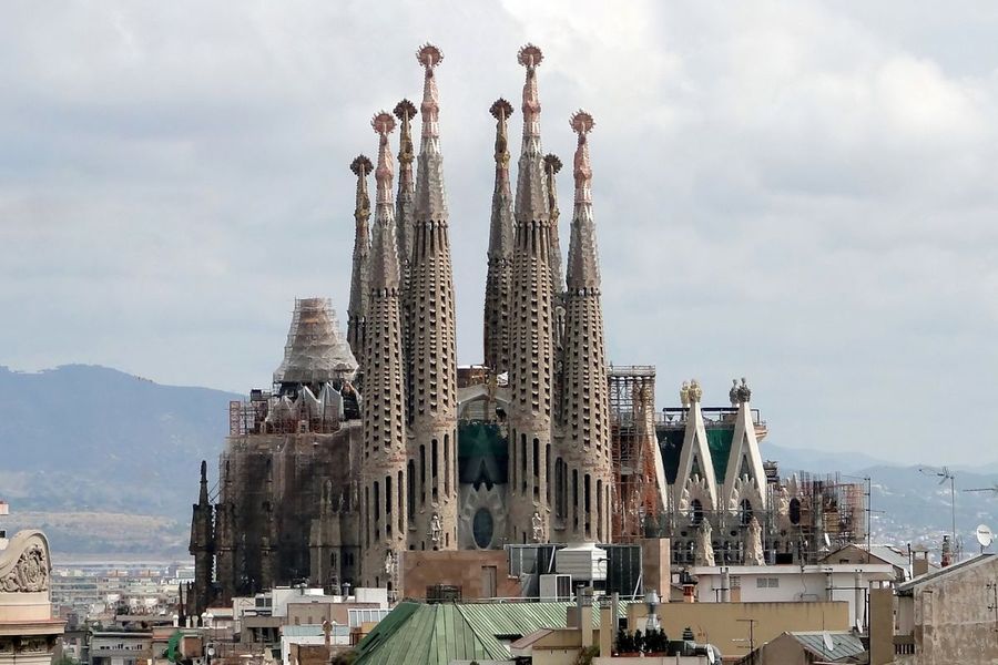 The stone spires of Barcelona's Sagrada Familia basilica rising above the surrounding rooftops under an overcast sky, with hills in the distance