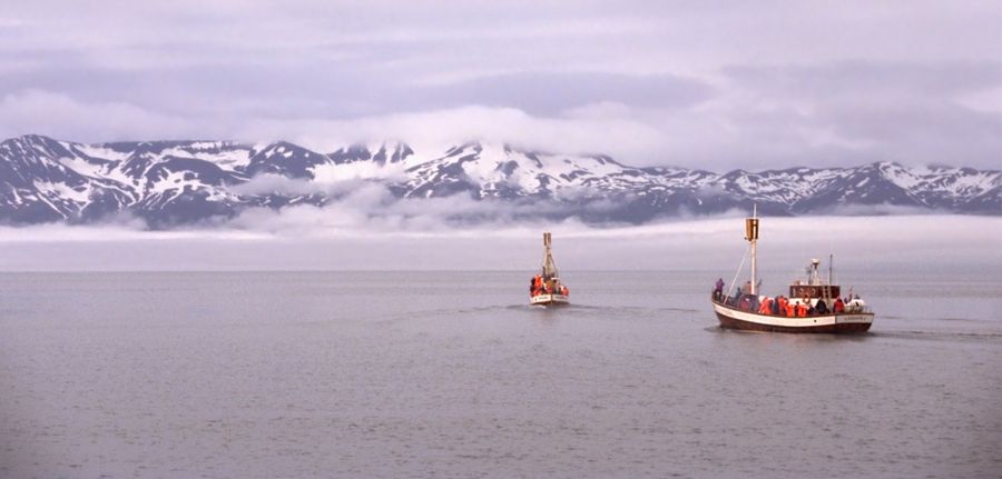Two traditional wooden oak whale-watching boats crossing Skjálfandi Bay from Húsavík, snow-streaked mountains rising behind low cloud
