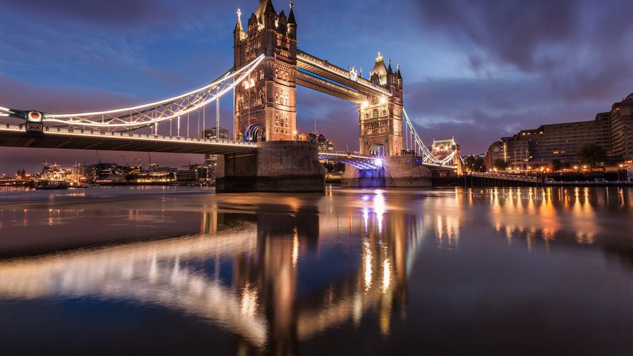 London's illuminated Tower Bridge reflected in the calm River Thames at dusk, with the lights of the city skyline glowing in the background