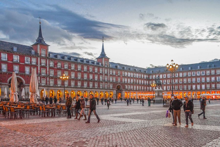 Madrid's arcaded Plaza Mayor at dusk, its red facades and twin spires lit up, with cafe tables to one side and people crossing the cobbled square