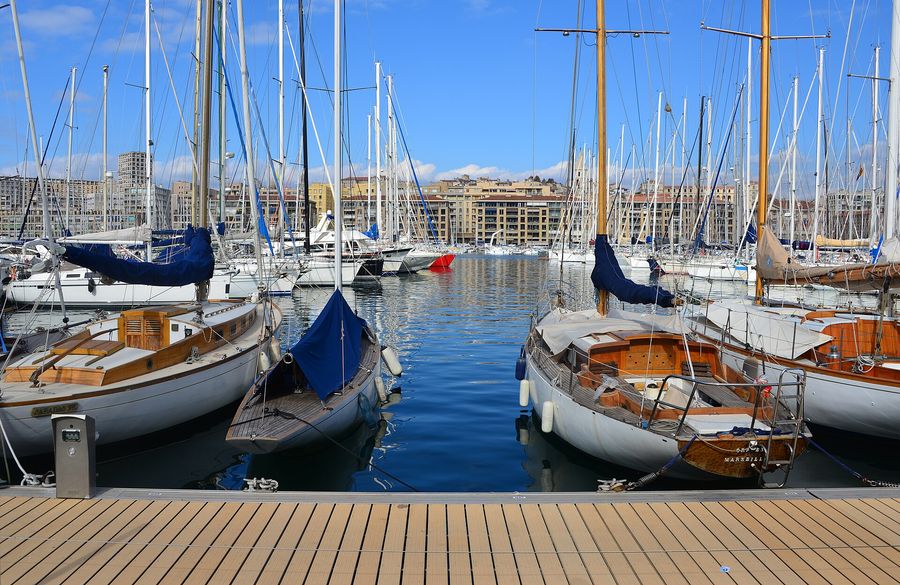 Classic wooden sailboats moored bow-to-quay in Marseille's Vieux-Port under a blue sky, with the city's pale apartment blocks and the hill of Notre-Dame de la Garde rising behind the harbour