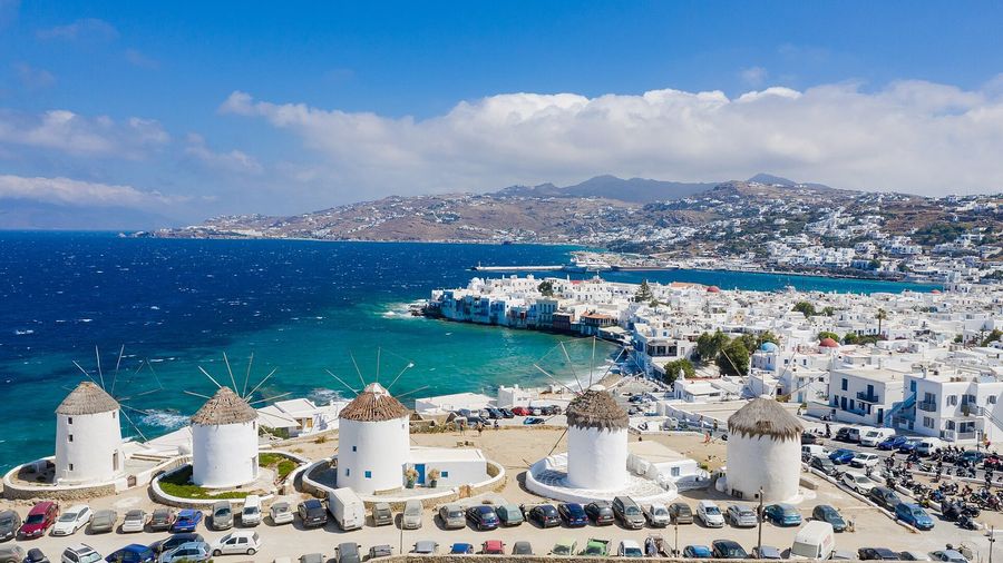 Aerial view of the row of whitewashed Kato Mili windmills above Mykonos Town, with the Little Venice waterfront, the harbour, and the turquoise Aegean bay beyond