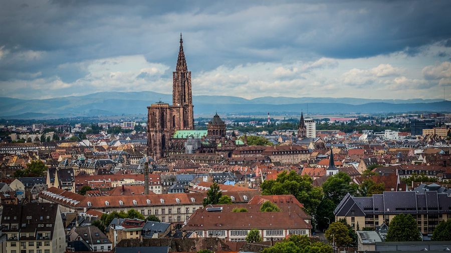Aerial view over the terracotta rooftops of Strasbourg's Grande Île toward the single pink-sandstone spire of the Gothic cathedral, with the Alsatian city stretching to the horizon