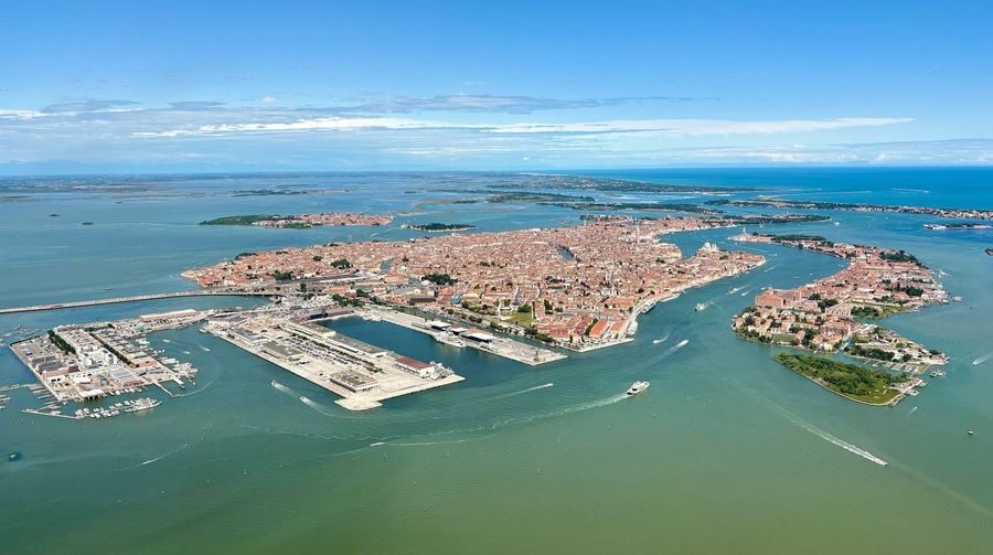 An aerial view of Venice and its dense terracotta rooftops rising from the shallow green lagoon, with outlying islands and the Adriatic Sea beyond