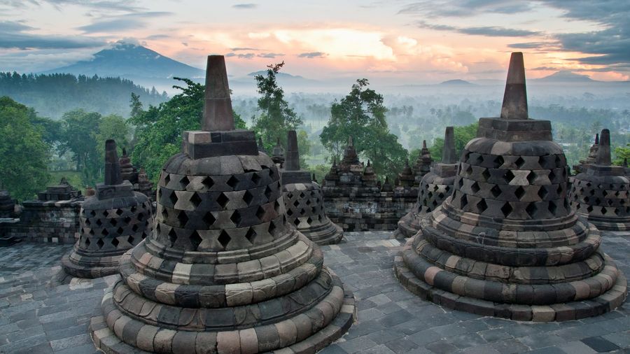 Dawn mist drifting over the stepped stupas and bell-shaped terraces of Borobudur temple near Yogyakarta, with volcanic hills silhouetted behind at sunrise