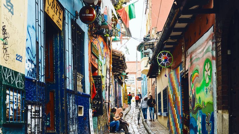 The colourful cobblestone Callejón del Embudo alley in Bogotá's La Candelaria, its colonial walls covered in murals and shop signs, with pedestrians and a cyclist heading uphill