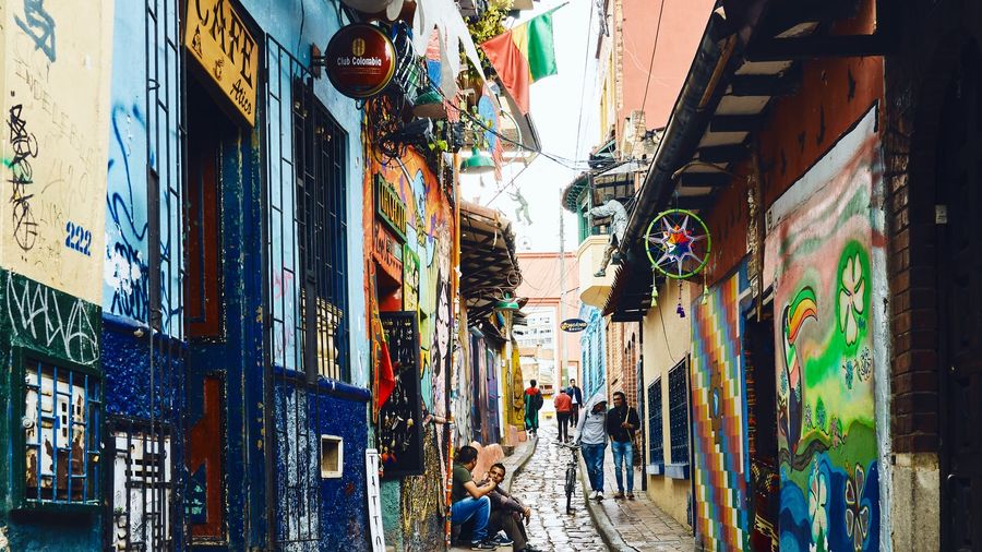 The colourful cobblestone Callejón del Embudo alley in Bogotá's La Candelaria, its colonial walls covered in murals and shop signs, with pedestrians and a cyclist heading uphill