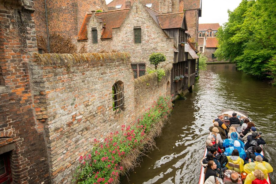A green canal tour boat of visitors gliding past medieval brick houses and a stone bridge on the reien of Bruges, Belgium