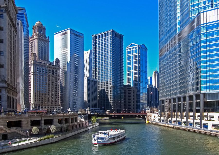 A white sightseeing tour boat cruising the Chicago River between downtown skyscrapers, passing beneath a bascule bridge