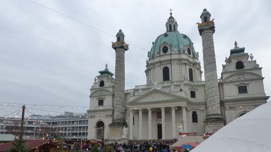 Wooden Christmas market stalls and strings of festive lights crowded with visitors in front of the domed Baroque Karlskirche in Vienna on an overcast winter day