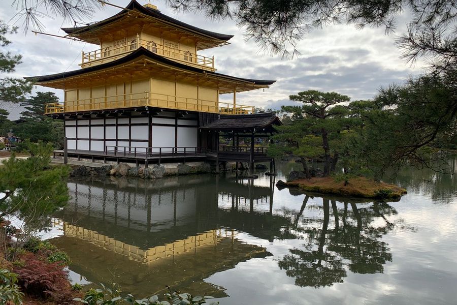 The gold-leaf-covered Kinkaku-ji Golden Pavilion in Kyoto mirrored in its still pond, framed by manicured pine trees under a cloudy sky