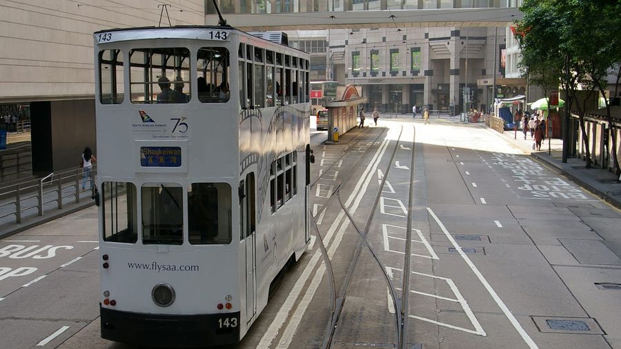 A white double-decker Hong Kong tram numbered 143 bound for Shaukeiwan, waiting at a tram stop on Des Voeux Road in Central beside office towers and a red minibus stand