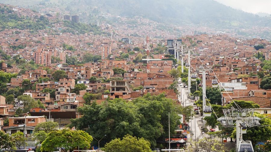 Metrocable gondola cabins climbing a cable line over the red-brick hillside barrios of Medellín, with a Metro station tower in the middle distance