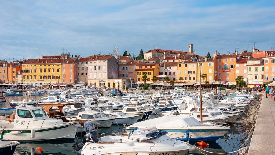 Small white boats packed into Rovinj's harbour below the pastel waterfront houses, with pedestrians on the quayside promenade on the Istrian coast