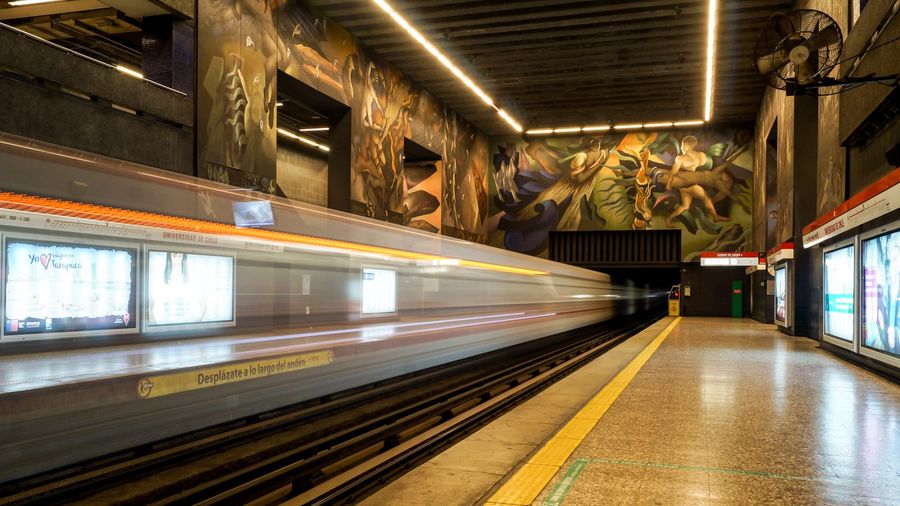 A Santiago Metro train blurs into Universidad de Chile station beneath Mario Toral's mural on Line 1 of the city's Red network
