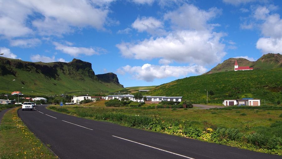 A car on the paved road into Vík í Mýrdal passing the village's white houses, with the red-roofed Reyniskirkja church on the green hillside and mountains rising beyond under a blue sky