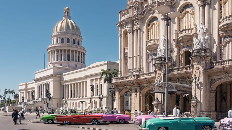 El Capitolio's floodlit neoclassical dome rising above the Paseo del Prado in central Havana at dusk, with brightly coloured 1950s American classic cars parked along the street in the foreground