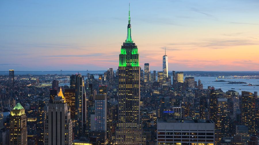 The Empire State Building rising above the illuminated Manhattan skyline at sunset, with One World Trade Center and New York Harbour in the distance