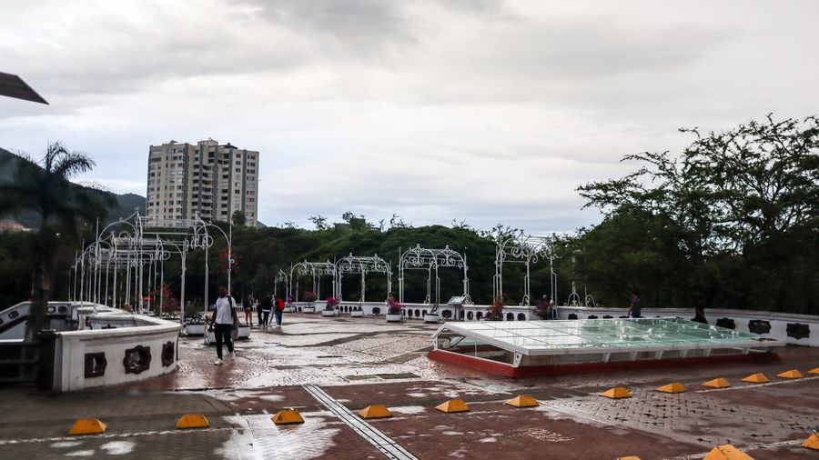 The Bulevar del Río pedestrian boulevard in downtown Cali, Colombia, with white wrought-iron arbours, flower planters, and the green hills of the Cauca Valley rising behind