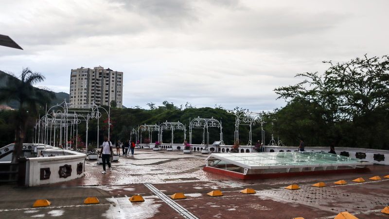 The Bulevar del Río pedestrian boulevard in downtown Cali, Colombia, with white wrought-iron arbours, flower planters, and the green hills of the Cauca Valley rising behind