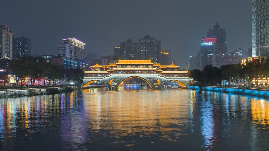 The illuminated Anshun covered bridge spanning the Jin River in Chengdu at night, its golden pavilion and lit arches mirrored in the water below the modern city skyline