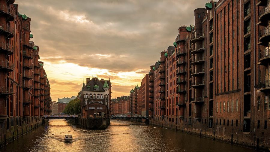 The moated Wasserschloss and red-brick warehouses of Hamburg's Speicherstadt district reflected in a canal at sunset, with a small boat passing beneath the iron bridges