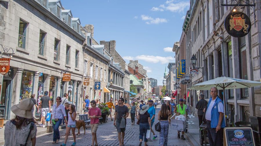 Pedestrians strolling the cobblestoned Rue Saint-Paul in Old Montreal on a sunny day, lined with historic grey-stone buildings, café terraces, and boutique signs