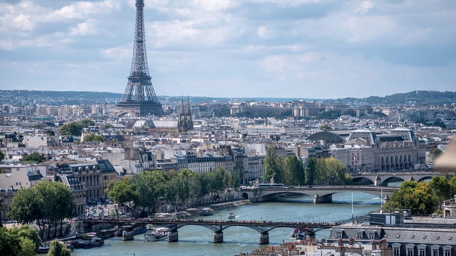 The Eiffel Tower rising above the rooftops of Paris with bridges spanning the River Seine in the foreground under a partly cloudy sky