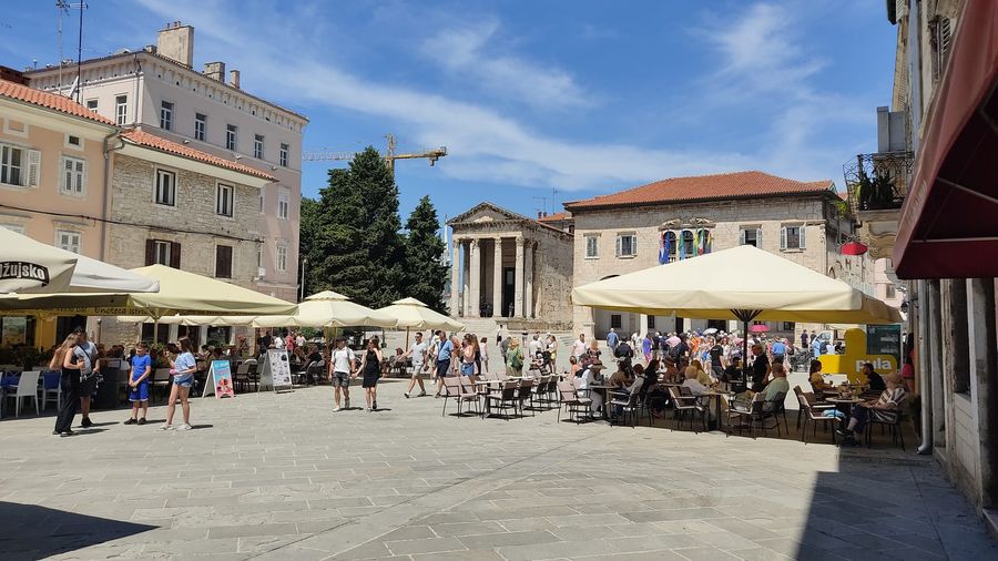 Pula's Roman Forum square with the Corinthian-columned Temple of Augustus behind café umbrellas and strolling visitors on a sunny day