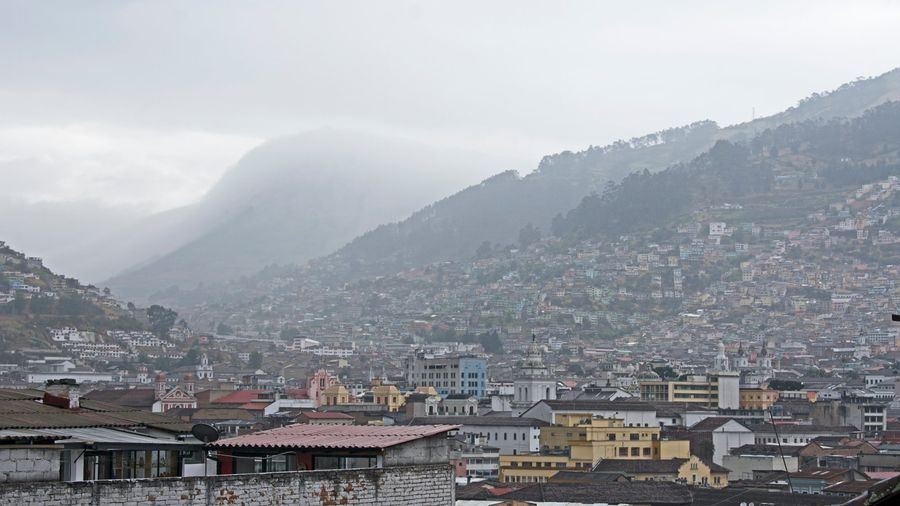 Panorama over the tiled roofs, church domes, and colonial towers of Quito's historic centre, with the Andean hills of the valley rising behind