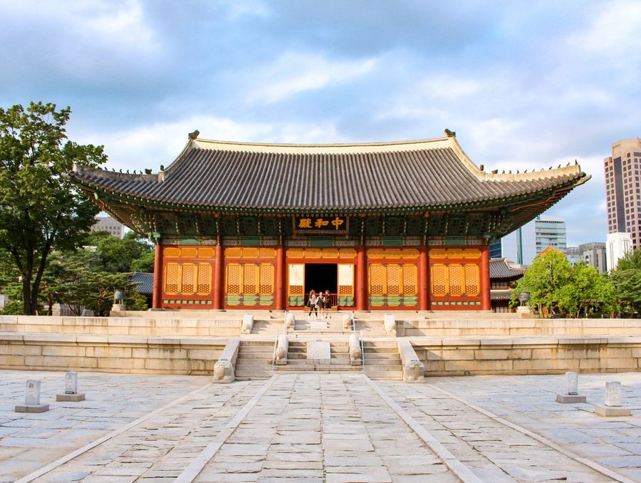 Junghwajeon, the red-and-gold wooden main throne hall of Deoksugung Palace in Seoul, set on a stone terrace with modern towers rising behind it