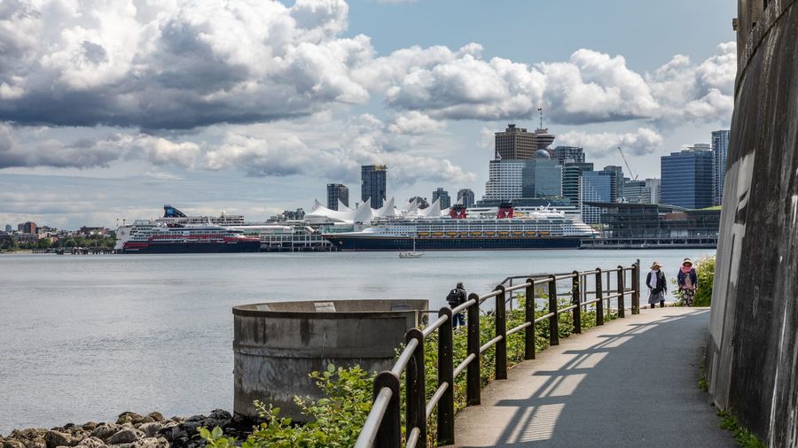 The Stanley Park seawall path curving along Vancouver's Coal Harbour, with walkers, moored cruise ships, the white sails of Canada Place, and the downtown skyline across the water