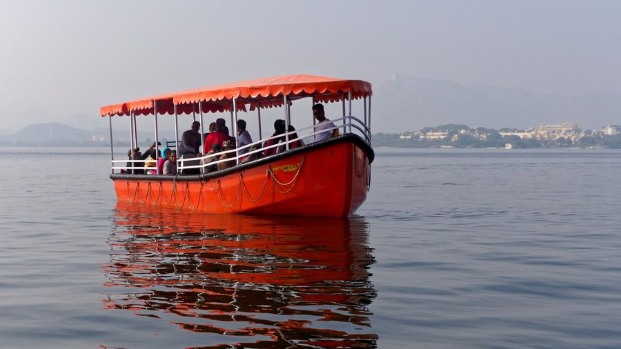 A wooden tourist boat crossing Lake Pichola in Udaipur, with the city's whitewashed lakeside buildings and the Aravalli hills rising behind the water