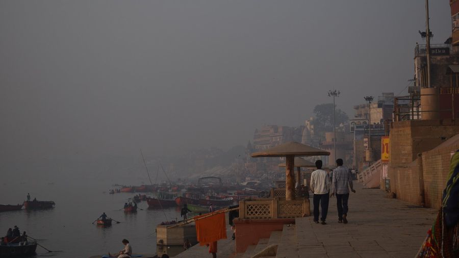 Wooden rowing boats moored and gliding along the misty Ganges at dawn while two people walk the stone ghat steps in Varanasi, India