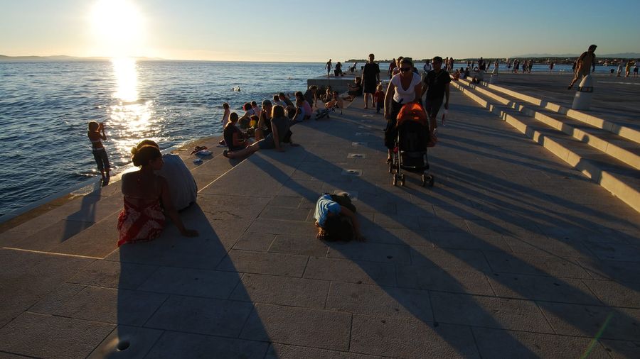 Visitors sitting on the marble steps of Zadar's Sea Organ at sunset, the wave-powered pipe holes visible in the pavement as the sun drops over the Adriatic islands